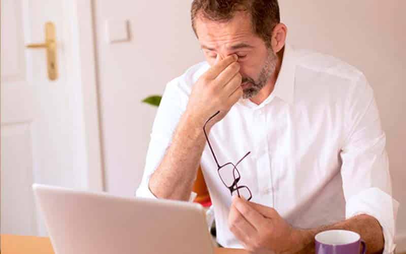 an exhausted man pinches the bridge of his nose with his eyes closed while at his laptop relating to home security mistakes to avoid